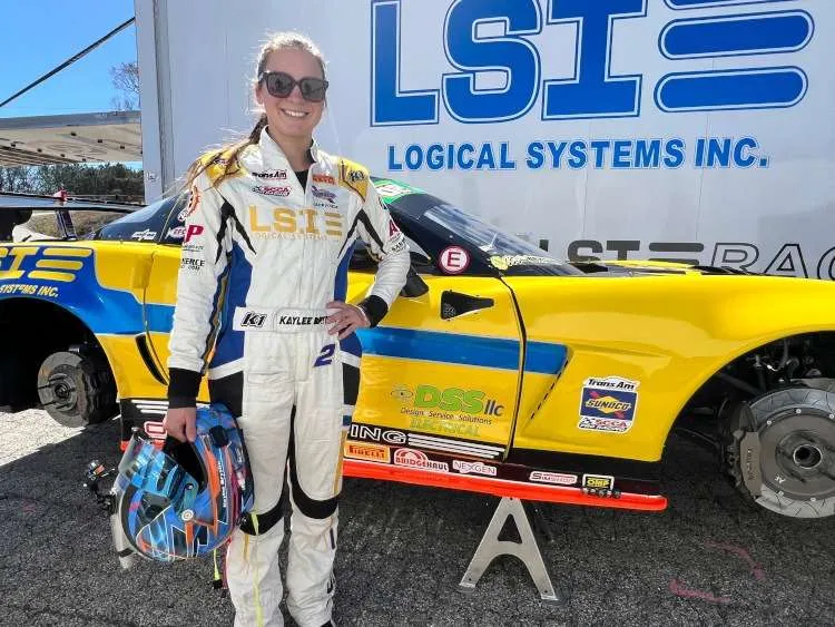 Kaylee Bryson, a talented race car driver for Logical Systems Inc., stands proudly beside her yellow and blue Corvette race car, which is propped up on jack stands.
