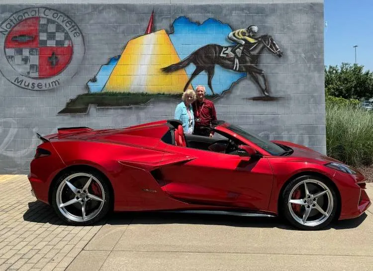 A radiant red 2024 Chevrolet Corvette E-Ray convertible is parked in front of a mural on the wall of the National Corvette Museum in Bowling Green, Kentucky. Two people, a woman in light blue and a man in red, stand smiling behind the car. The mural depicts the National Corvette Museum logo, an outline of Kentucky with a horse racer, and other elements.
