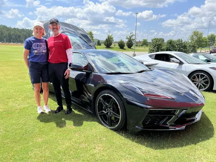 "Etta and Glen proudly standing next to their black 2024 C8 Corvette coupe with the engine compartment open, displayed at an outdoor car show in Cartersville, Georgia."