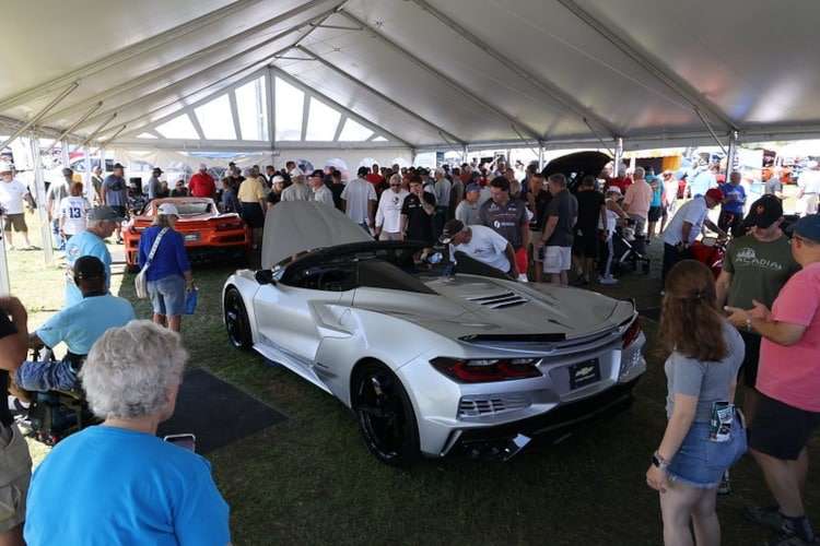 A silver Chevrolet C8 Corvette convertible with its rear engine cover open, displayed under a tent with a large crowd of spectators at the Corvettes at Carlisle 2025 event. Photo from Vettes of Atlanta Magazine.