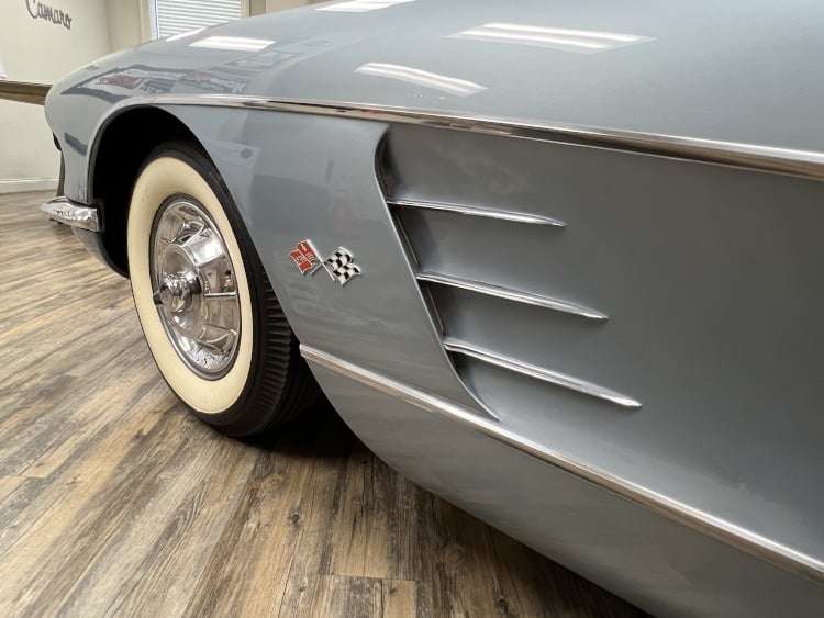 Close-up of the front fender and side cove on the driver's side of a silver 1958 Corvette, featuring the classic chrome trim and the crossed-flags emblem.