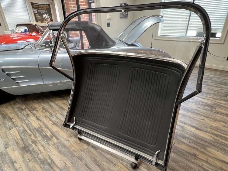 A removable hard top for a 1958 Corvette stands upright, showing the black interior and chrome trim, with the car it belongs to visible in the background.
