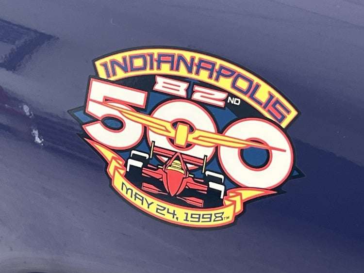 A close-up shot of the official sticker logo for the 82nd Indianapolis 500, placed on the purple body of a 1998 Chevrolet Corvette C5 Pace Car. The logo features a red IndyCar race car over a yellow banner with "82nd" and "Indianapolis 500" text, and the date "May 24, 1998" on a lower banner. The photo is from Vettes of Atlanta Magazine.