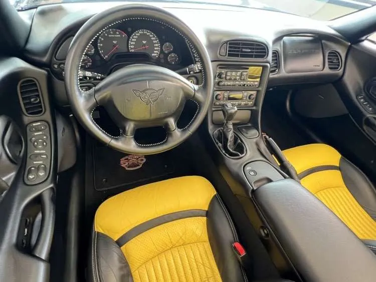 A professional photograph of the driver's side interior of the 1998 Chevrolet Corvette C5 Indy 500 Pace Car. The image shows the black and yellow leather bucket seat, the black leather-wrapped steering wheel with white stitching, and the dashboard featuring the instrument cluster and center console. A floor mat with the 'Indianapolis 500' logo is visible. Photo credit: Vettes of Atlanta Magazine.