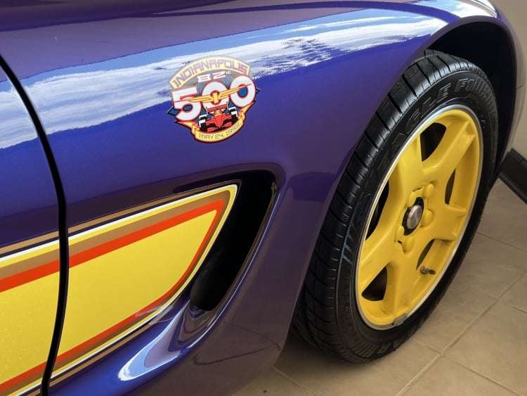 A close-up, angled photograph showing the side of the 1998 Chevrolet Corvette C5 Indy 500 Pace Car. The image focuses on the front fender, highlighting the official Indianapolis 500 logo decal with the words "Indianapolis 500" and "82nd" along with an illustration of a race car. The car's blue paint reflects the surroundings, and a yellow and red accent stripe runs along the side. The matching yellow five-spoke wheel and part of the tire are also visible. The image is a professional photograph owned by Vettes of Atlanta Magazine.