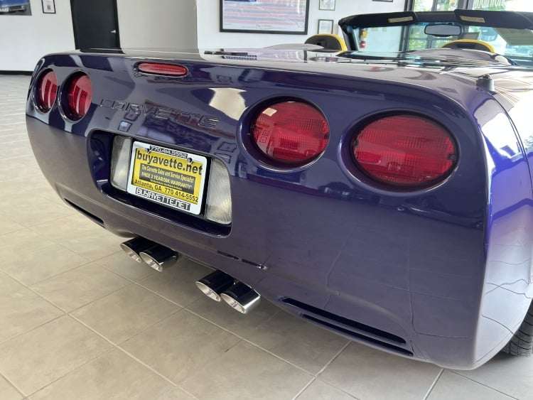A rear-quarter view photograph of a 1998 Chevrolet Corvette C5 Indy 500 Pace Car. The image focuses on the rear of the car, highlighting the iconic four circular red taillights and the central "CORVETTE" embossed lettering on the bumper. The license plate frame advertises "buyavette.net" and provides contact information for a dealership in Atlanta, GA. The car's blue metallic paint and four chrome exhaust tips are also visible. The image is a professional photograph owned by Vettes of Atlanta Magazine.