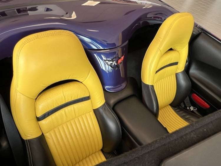 A high-angle close-up photograph of the interior of a 1998 Chevrolet Corvette C5 Indy 500 Pace Car convertible. The image focuses on the two-tone black and yellow leather bucket seats and the "waterfall" between them. The waterfall, painted in the car's exterior blue color, features the special edition '82nd Indianapolis 500' logo on its side. The convertible's dark purple hard tonneau cover is partially visible, and the overall interior has a clean, polished look. The image is a professional photograph owned by Vettes of Atlanta Magazine.