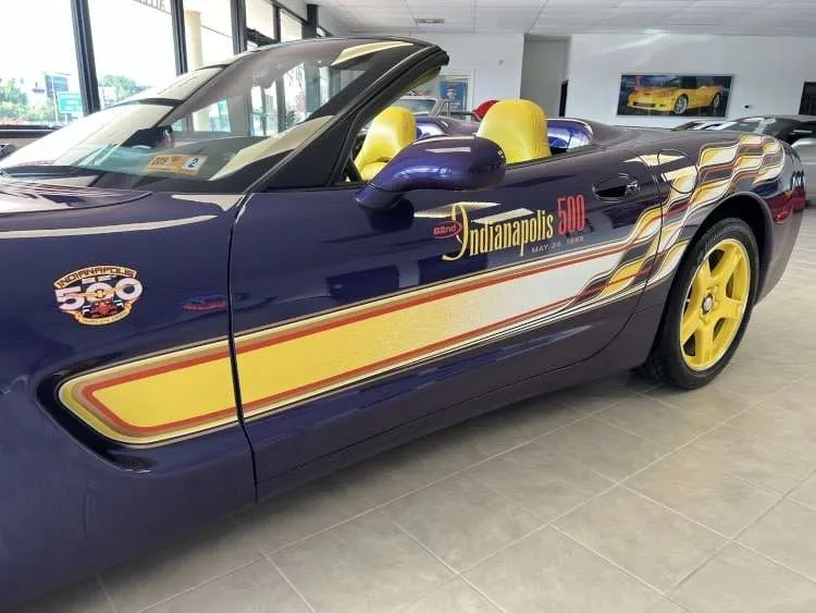 A side view of a rare 1998 Chevrolet Corvette C5 convertible, the official Indianapolis 500 Pace Car. The car is painted in a distinctive Radar Blue with bold yellow accents, including the seats, wheels, and a graphic stripe that features the "82nd Indianapolis 500 May 24, 1998" logo on the door. The image, captured in a showroom, highlights the iconic design and unique colors of this limited-edition model.