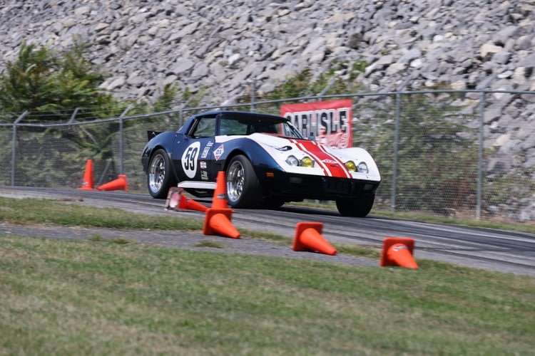 Photo of a classic blue and white C3 Corvette race car, number 59, navigating an autocross course at Corvettes at Carlisle 2025 with orange cones marking the autocross track.