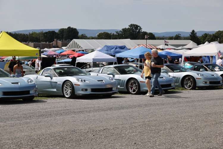 A long row of Carlisle Blue Corvettes parked at Corvettes at Carlisle 2025. People are walking between the cars and vendor tents are visible in the background.