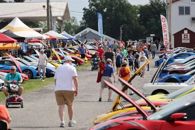 Photo showing a large row of new and classic Corvettes with their hoods open on the 'Fun Field' at Corvettes at Carlisle 2025, as a crowd of people walks and browses the vehicles.