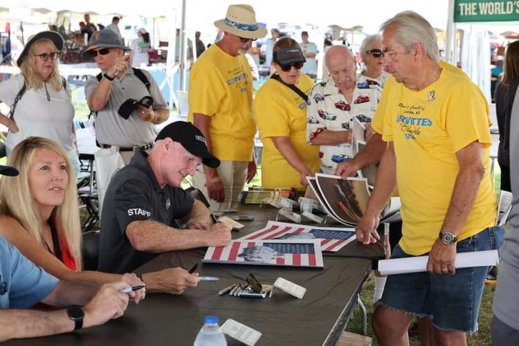 Photo of a celebrity or guest at Corvettes at Carlisle 2025, a 'Staff' member in a black shirt, signing autographs and interacting with a crowd of attendees in yellow event shir
