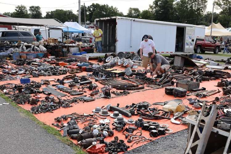 A panoramic view of the bustling swap meet at Corvettes at Carlisle 2025, captured for Vettes of Atlanta Magazine, showing a vast array of vintage Corvette parts on orange tarps, with enthusiasts browsing and vendor trailers in the background.