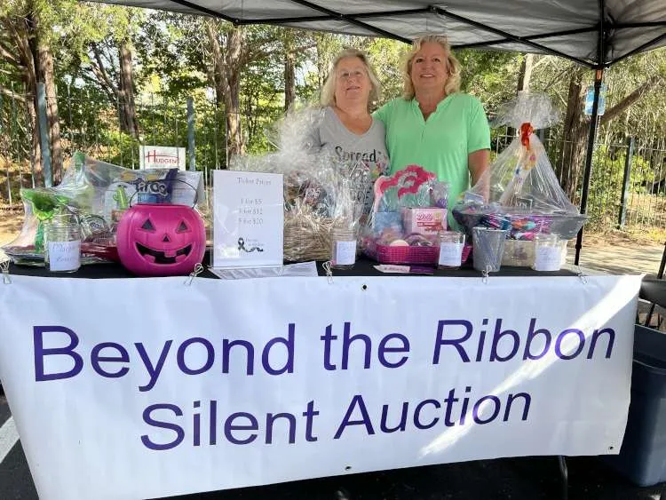 Two smiling women stand behind a table laden with silent auction items and a "Beyond the Ribbon Silent Auction" banner, featuring gift baskets, a pink pumpkin, and ticket prices.