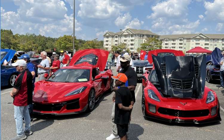 Two striking red Corvettes, one a newer mid-engine model with its door open and the other an earlier model with its hood up, are prominently featured at an outdoor car show. Spectators, including a man and a child in the foreground, admire the vehicles under a partly cloudy sky. Residential buildings are visible in the distant background.