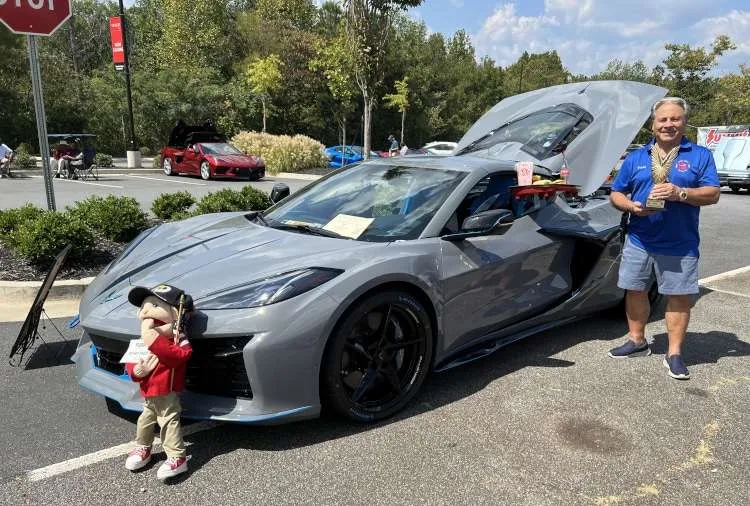 A man in a blue shirt and shorts stands next to a gray Chevrolet Corvette, holding a trophy and smiling. The car's trunk is open, revealing a small tray with snacks. A puppet figure stands in front of the car, adding a playful touch. The scene is an outdoor car show under a sunny sky, with other cars and trees in the background.