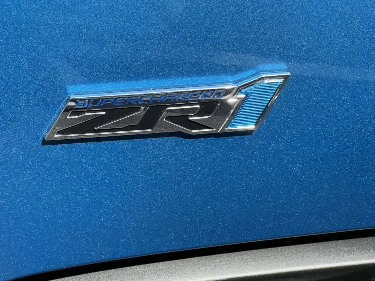 Alt-Text: Close-up of the chrome and blue "Supercharged ZR1" emblem on the exterior fender of a blue metallic 2009 Chevrolet Corvette.