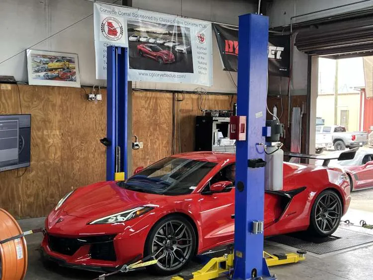 A Torch Red Corvette C8 Z06, with its high-revving mid-engine platform, is precisely centered and secured on the Dynojet rollers at Marshall’s Performance. The supercar is framed by vibrant blue lift posts and features a prominent rear wing. In the background, banners for the Classic Glass Corvette Club and the National Corvette Museum hang above the technical workstation, while a red C7 Z06 waits outside the open bay door.