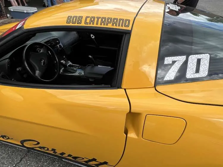 Close-up of the driver-side door and roofline of a yellow Chevrolet Corvette C6, featuring "BOB CATAPANO" name decal on the roof rail and racing number 70 on the rear quarter window.