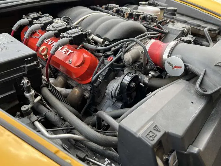 Close-up view of a high-performance Chevrolet Corvette C6 LS V8 engine bay, highlighting a set of custom red 'Chevrolet' branded valve covers, red spark plug wires, a detailed alternator, power steering reservoir, and custom air intake system.