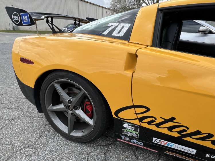 Passenger-side rear quarter view of a yellow Chevrolet Corvette C6 track car, showing the high-mount racing wing, charcoal five-spoke wheels, red brake calipers, and racing number 70 on the window.