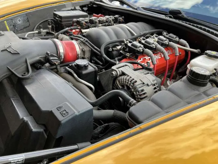 Close-up of a Chevrolet Corvette C6 engine bay, featuring a high-performance LS engine with custom red Chevrolet valve covers, a detailed alternator, power steering reservoir, and stock air intake system.