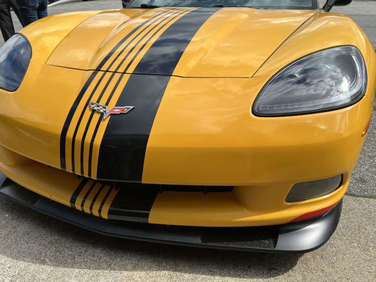 Close-up front view of a yellow Chevrolet Corvette C6, highlighting the large black multi-stripe racing decal, the classic crossed-flags emblem, a modern LED-style headlight, and a black front splitter with a small red accent.