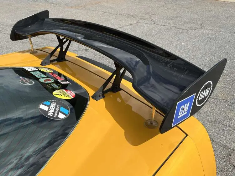 High-angle close-up of a gloss black racing wing spoiler mounted on a yellow Chevrolet Corvette, featuring GM and UAW decals on the endplates and track-day stickers on the rear window.