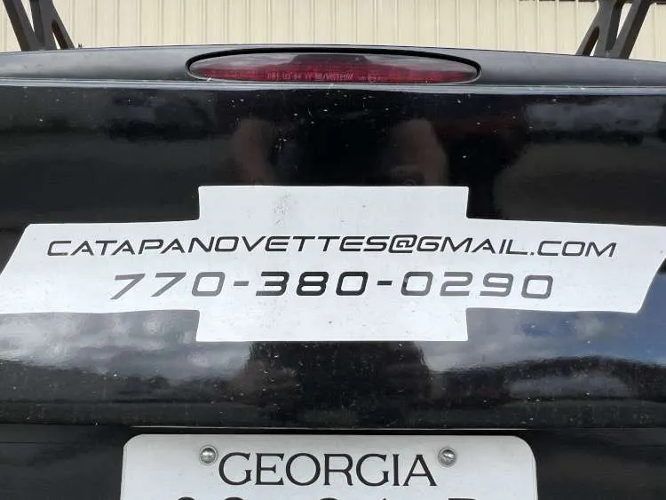 Close-up of a black Chevrolet Corvette rear bumper with a silver bow tie decal featuring contact details: "CATAPANOVETTES@GMAIL.COM" and phone number "770-380-0290", above a partial Georgia license plate and the center high-mount stop light.