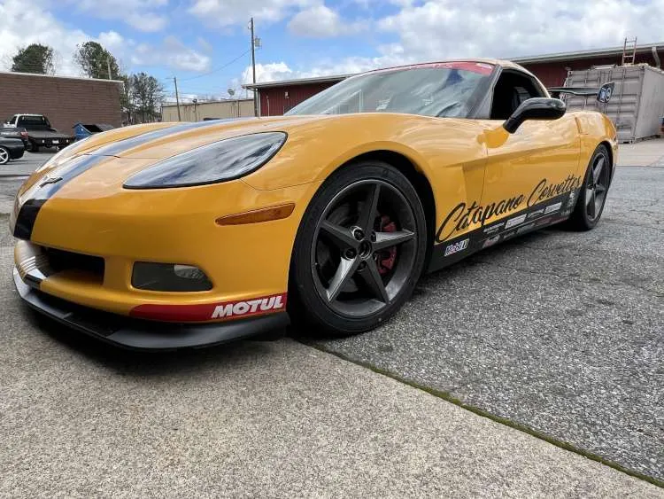 Low-angle wide-angle shot of a yellow Chevrolet Corvette C6 track car with black racing stripes, Catapano Corvettes door decals, Motul front splitter branding, and a high-mount rear wing.