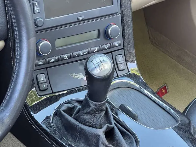 A detailed close-up photograph of a customized silver short-throw manual shifter in a modified Chevrolet Corvette C6, featuring a carbon fiber center console overlay, a round metallic gear knob, leather boot, and the traction control button.