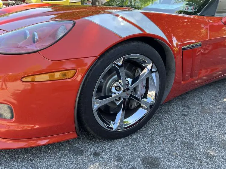 Driver-side front angle of a metallic orange C6 Corvette Grand Sport, showing the signature fender vents, aftermarket black multi-spoke wheels, carbon fiber side mirrors, and front splitter.