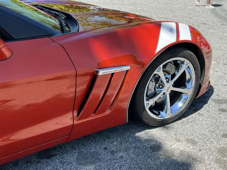Detailed low-angle front passenger-side view of a custom dark metallic blue C7 Chevrolet Corvette Z06. The image highlights its extensive carbon fiber aero kit (front splitter, side skirts, fender vents, mirror covers) and custom black/silver multi-spoke alloy wheels with prominent "Corvette" script brake calipers.