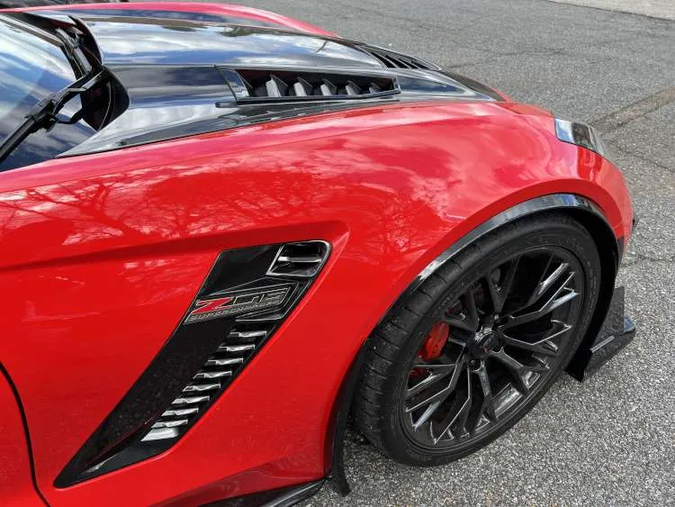Close-up of a red C7 Corvette Z06 front passenger fender featuring the black Z06 Supercharged emblem, side vents, and carbon fiber hood accents.