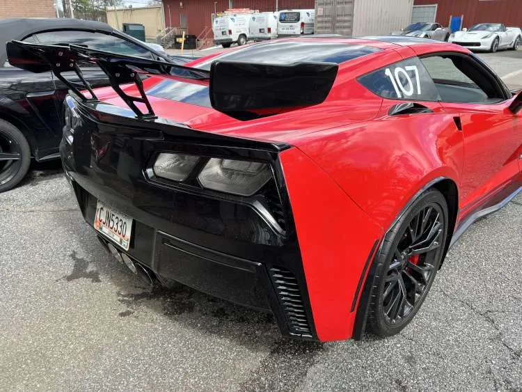 Rear three-quarter view of a red C7 Corvette Z06 featuring a large black high-wing racing spoiler, clear taillights, and track number 107 on the window.