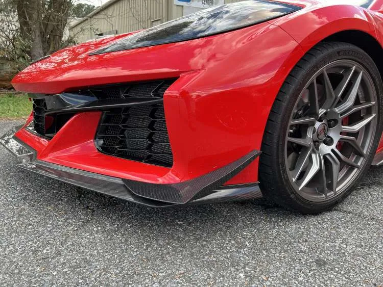 Low-angle view of a red C8 Corvette Z06 front bumper featuring a carbon fiber splitter with dive planes, Michelin Pilot Sport Cup 2 R tires, and open radiator intakes.