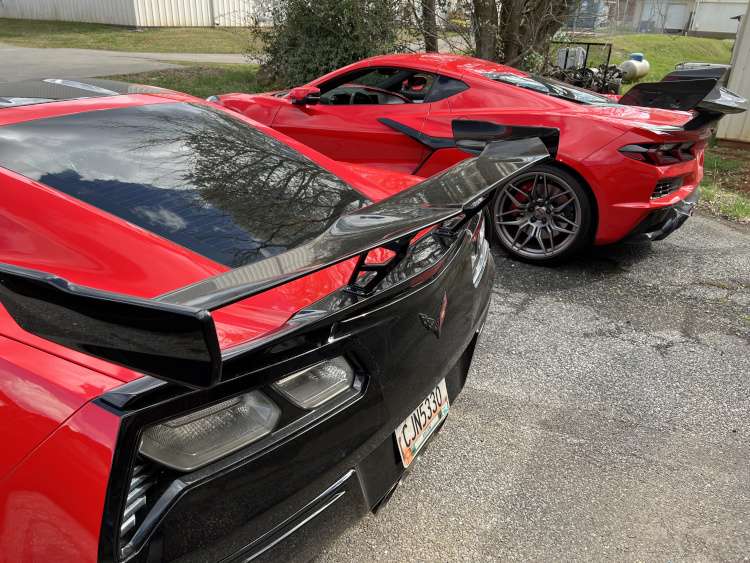 Rear view comparison of a red C7 Corvette Z06 and a red C8 Corvette Z06, both featuring large carbon fiber high-wing spoilers and performance aero kits.