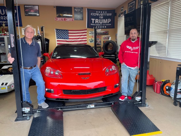 Two men in a garage beside a sixth-generation red Corvette