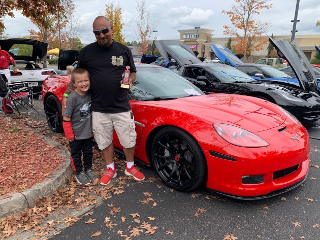 Father and son holding a car show award beside a Corvette