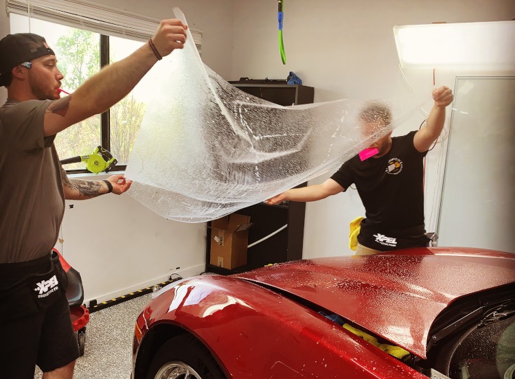 Two guys installing XPEL ppf on the hood of red Corvette