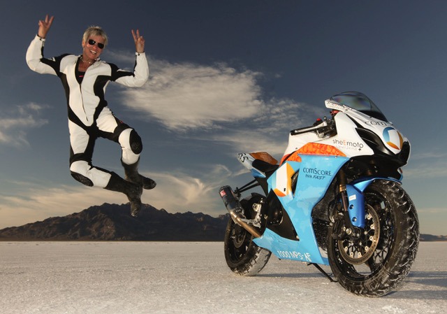 A women in racing leather jumping beside a motorcycle