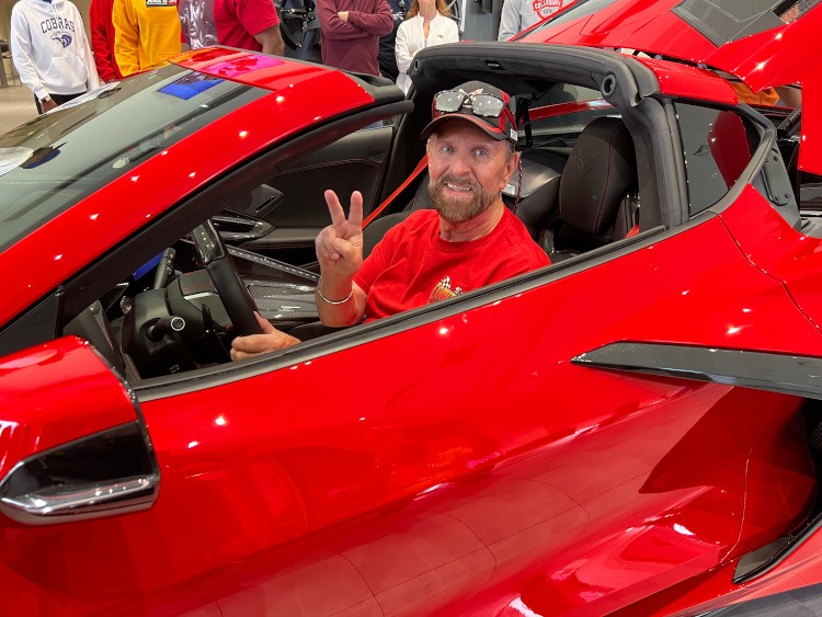 Man flashing peace sign in a torch red C8 Z06 Corvette coupe