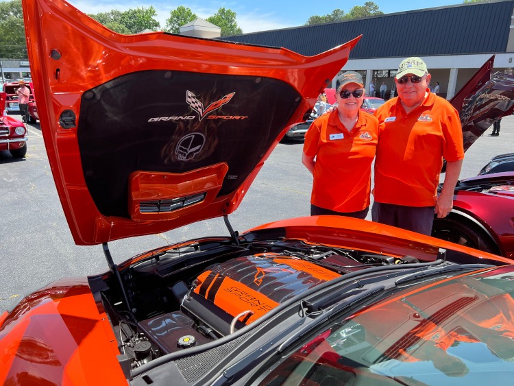Two people are standing beside a Sebring Orange Corvette coupe.