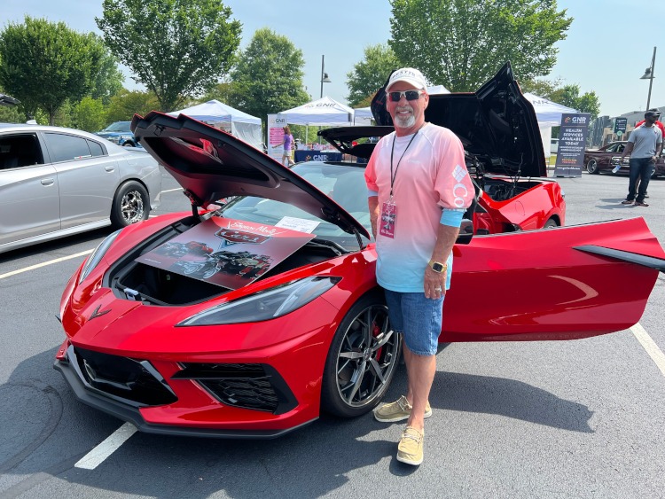 A man is standing beside a Torch Red Corvette convertible.