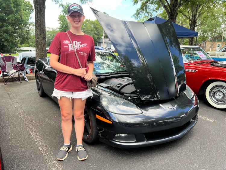 A girl stands beside a C6 black Corvette coupe.