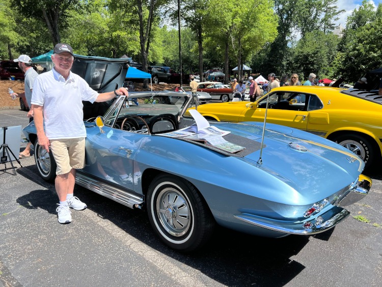 A man stands beside a 1964 blue Corvette convertible at a car show.