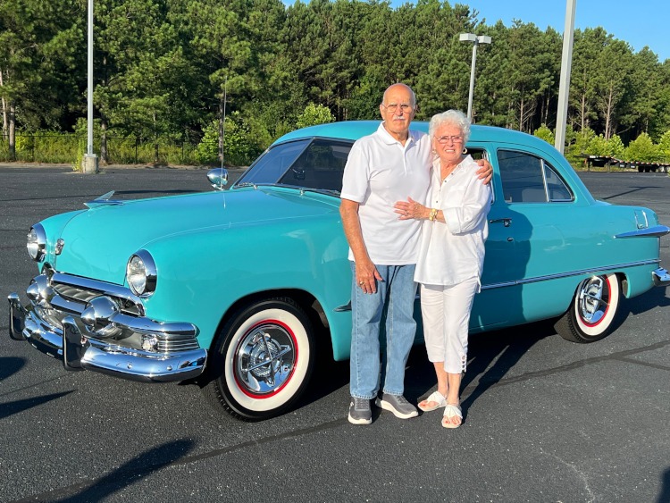 People are standing beside a 1951 teal-colored vintage Ford car.