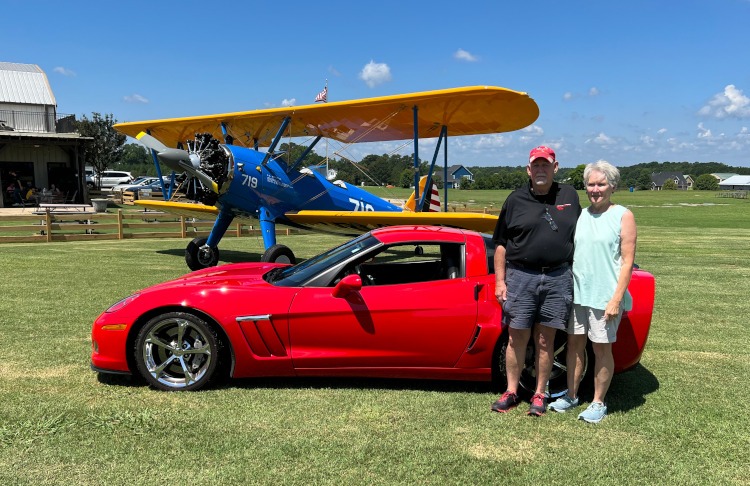 Two people are standing beside a 2011 Grand Sport Corvette with a bi-plane in the background.