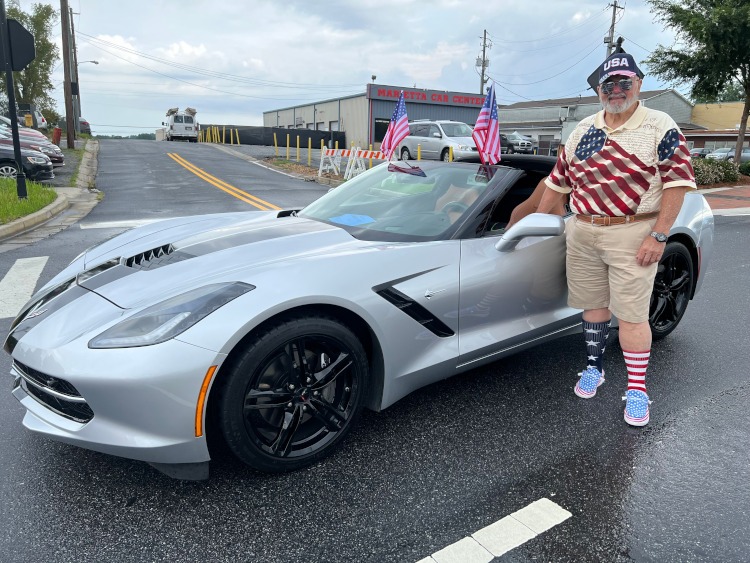 A man stands beside a grey Corvette coupe at a 4th of July parade.
