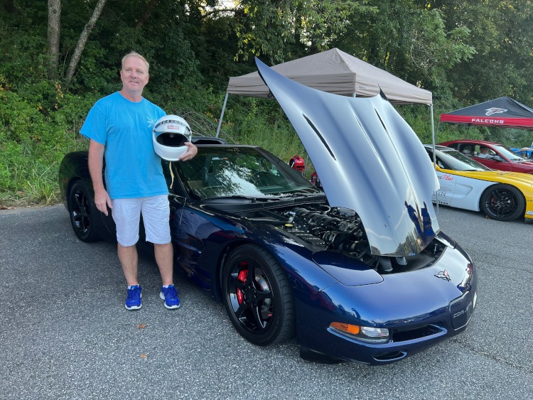 A man stands beside a fifth-generation blue Corvette coupe with his helmet.